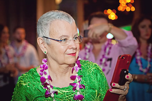 Picture of La Rita Mason: a woman facing to the right, with short gray hair wearing a green patterned shirt and a lei of purple flowers. She is holding a red phone.